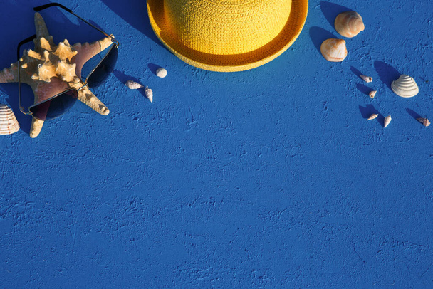 Frame with beach accessories on a nautical theme: yellow straw hat, sunglasses, starfish and shells on a blue background. Vacation concept, sea trip, UV protection, swimming. Copy space. Flatlay - Fotó, kép