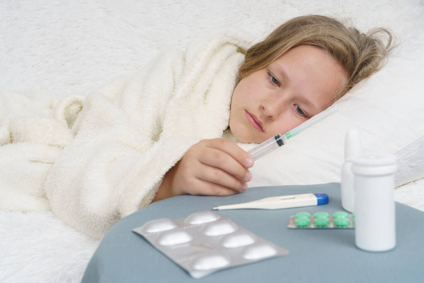 Μια άρρωστη έφηβη ξαπλώνει στο κρεβάτι και κρατά μια σύριγγα στα χέρια της, δίπλα της είναι ένα κομοδίνο με φάρμακα και ένα θερμόμετρο. Ιατρική έννοια. - Φωτογραφία, εικόνα