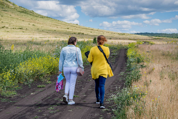 Две девушки в летней одежде поднимаются на вершину холма вдоль грунтовой дороги вдоль зеленого поля. - Фото, изображение