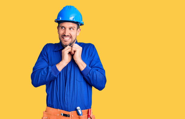 Junger gutaussehender Mann in Arbeiteruniform und Hut, der nervös und aufgeregt lacht, die Hände am Kinn zur Seite gerichtet  - Foto, Bild