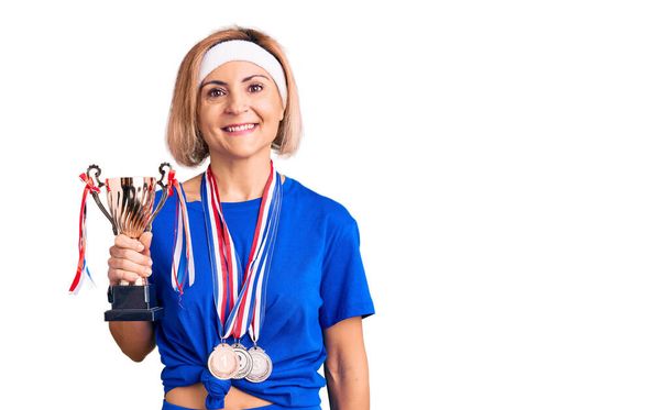 jonge blonde vrouw houden kampioen trofee dragen medailles kijken positief en gelukkig staan en glimlachen met een zelfverzekerde glimlach tonen tanden  - Foto, afbeelding