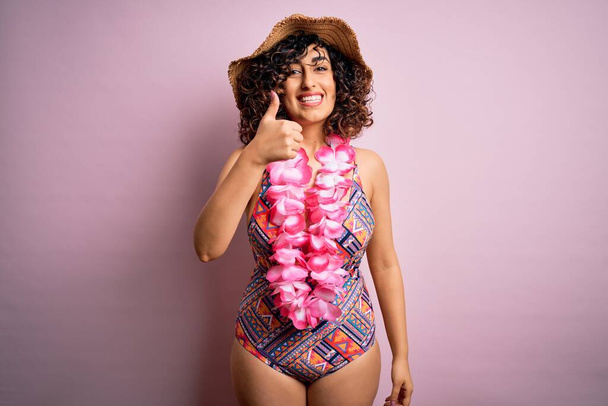 Jonge mooie arabische vrouw op vakantie in badpak en Hawaïaanse lei bloemen doet vrolijke duimen omhoog gebaar met de hand. Goedkeurende uitdrukking die naar de camera kijkt en succes laat zien. - Foto, afbeelding