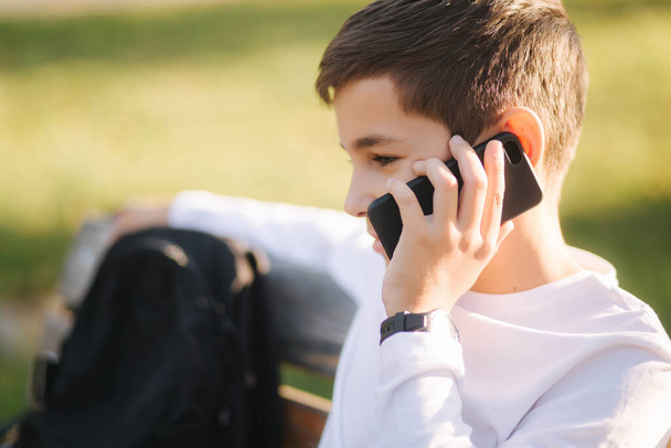 Young boy in white sweatshirt with black backpack sitting on the bench in the park and speak with somebody by the phone - Foto, imagen