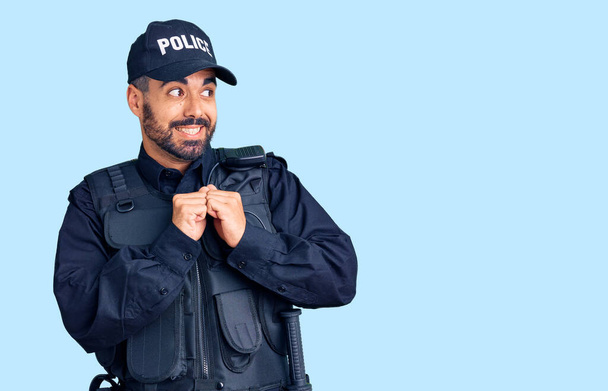 Junger hispanischer Mann in Polizeiuniform lacht nervös und aufgeregt, die Hände am Kinn zur Seite gerichtet  - Foto, Bild