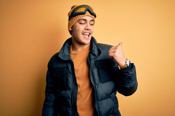 Fiatal brazil síelő férfi visel hó sportruházat és síszemüveg sárga háttér mosolyog boldog arccal néz, és rámutatva oldalra hüvelykujjával felfelé. - Fotó, kép