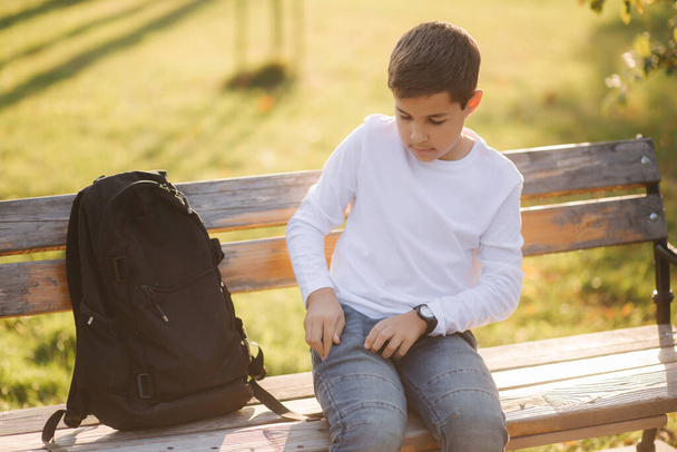 Jonge jongen haalt zijn zaktelefoon uit zijn zak. Tienerjongen zittend op de bank in het park - Foto, afbeelding