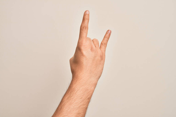 Χέρι του καυκάσιου νέου άνδρα που δείχνει τα δάχτυλά του πάνω από απομονωμένο λευκό φόντο gesturing ροκ εν ρολ σύμβολο, δείχνοντας άσεμνη χειρονομία κέρατα - Φωτογραφία, εικόνα