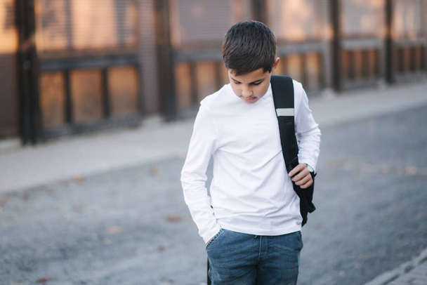 A tinédzser fiú kivesz egy vezeték nélküli fejhallgatót, és beteszi a fülébe. Fiatal fiú fehér ingben, hátizsákkal. Hallgasd a zenét. - Fotó, kép