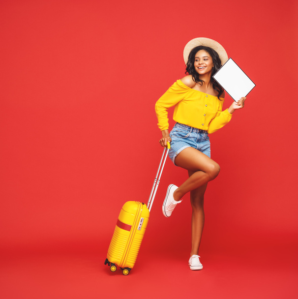 turista étnico feminino otimista de comprimento total com mala e cartaz em branco sorrindo e olhando para longe contra backdro vermelho
 - Foto, Imagem