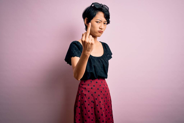 Молодая красивая азиатская девушка в повседневном платье, стоящая на изолированном розовом фоне, демонстрируя средний палец, невежливый и грубый отъебать выражение лица
 - Фото, изображение