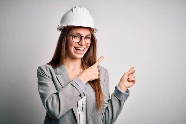 Fiatal gyönyörű vörös hajú építész nő visel biztonsági sisak felett fehér háttér mosolyog, és néz a kamera mutat két kézzel és ujjal oldalra. - Fotó, kép