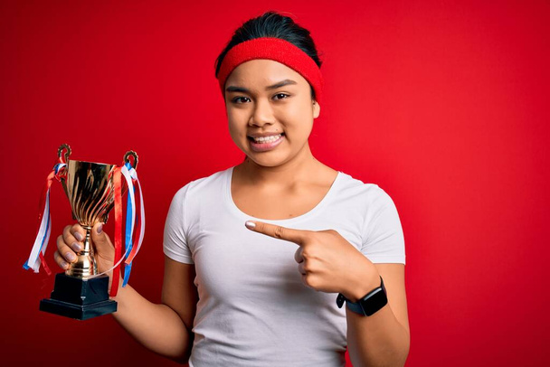 Joven campeón asiático chica ganando trofeo taza de pie sobre aislado rojo fondo muy feliz señalando con la mano y el dedo
 - Foto, Imagen