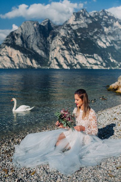 a girl in a smart white dress is sitting on the embankment of lake Garda.A woman is photographed against the background of a mountain and lake in Italy.Torbole. - Photo, image