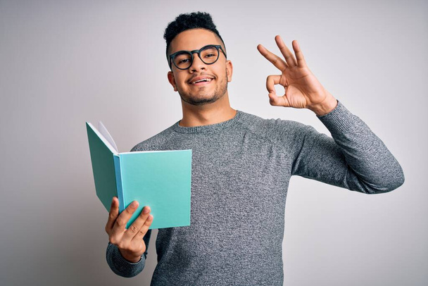 Joven estudiante inteligente guapo leyendo libro sobre fondo blanco aislado haciendo signo ok con los dedos, símbolo excelente
 - Foto, imagen