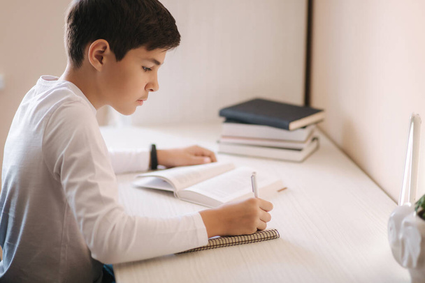 Młody chłopiec siedzący za biurkiem czyta książkę i zapisuje w notatniku. Badanie w domu podczas kwarantanny - Zdjęcie, obraz