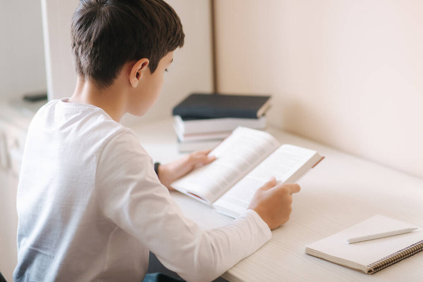 Ein kleiner Junge, der am Schreibtisch sitzt, liest das Buch und blättert in einem Notizbuch. Während der Quarantäne zu Hause studieren - Foto, Bild