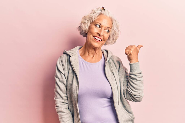 Starsza siwowłosa kobieta ubrana w luźne sportowe ubrania uśmiechnięta ze szczęśliwym wyglądem twarzy i wskazująca na bok z kciukiem w górze.  - Zdjęcie, obraz