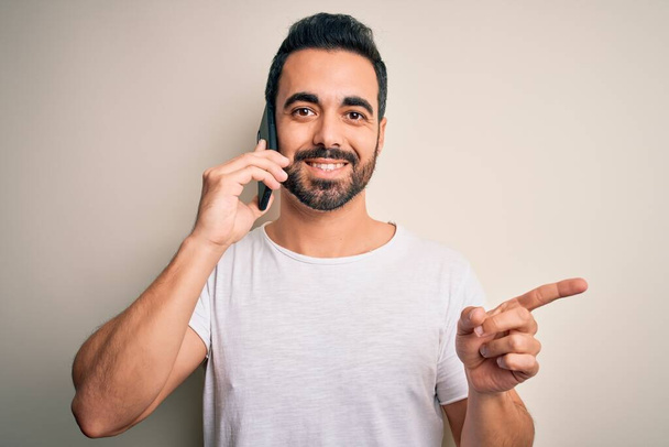 Nuori komea mies, jolla on parta, jolla on keskustelu älypuhelimessa, erittäin onnellinen osoittaen kädellä ja sormella sivulle - Valokuva, kuva