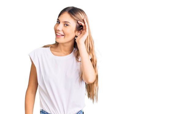 Beautiful caucasian woman with blonde hair wearing casual white tshirt smiling with hand over ear listening an hearing to rumor or gossip. deafness concept.  - Photo, Image