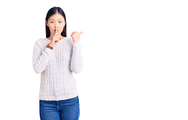 Młoda piękna Chinka ubrana w luźny sweter, prosząca o ciszę z palcem na ustach wskazującym ręką na bok. cisza i tajne pojęcie.  - Zdjęcie, obraz