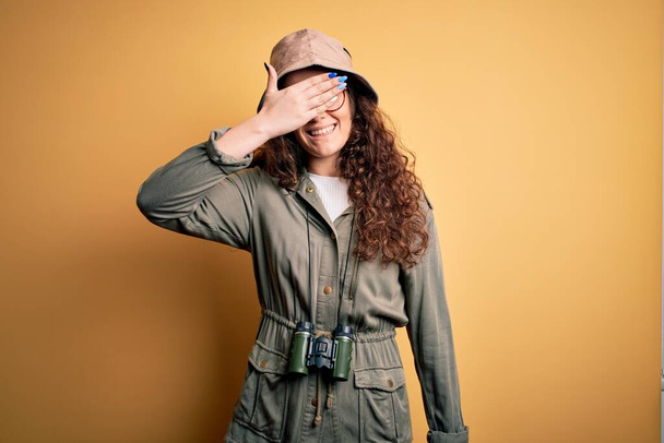 Mladá krásná turistka na dovolené v průzkumné čepici a dalekohledu s úsměvem a smíchem s rukou na tváři zakrývající oči pro překvapení. Slepý koncept. - Fotografie, Obrázek