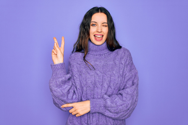 Молодая красивая женщина в свитере в водолазке стоит на фиолетовом фоне улыбаясь счастливым лицом, подмигивая в камеру, делая знак победы пальцами. Номер два:
. - Фото, изображение