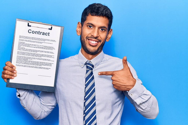 Jeune homme latin tenant presse-papiers avec document contractuel pointant du doigt un sourire heureux et fier  - Photo, image