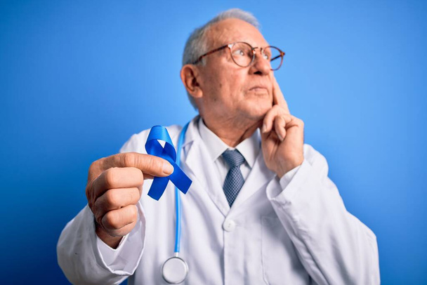 Siwy starszy lekarz trzymający świadomość raka okrężnicy Niebieska wstążka na niebieskim tle poważna twarz myśląca o pytaniu, bardzo zdezorientowany pomysł - Zdjęcie, obraz