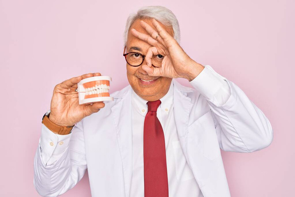 Середнього віку старший сірошерстий стоматолог тримає протезний зуб на рожевому фоні з щасливим обличчям, посміхаючись, роблячи знак ок з рукою на очах, дивлячись через пальці
 - Фото, зображення