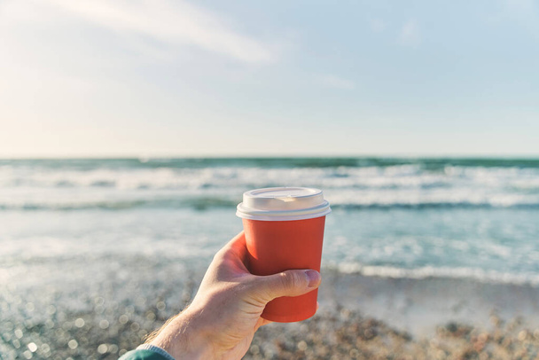 A young man holds a paper cup with coffee on the background of the sea - 写真・画像