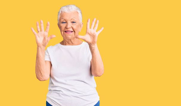 Ältere schöne Frau mit blauen Augen und grauen Haaren trägt legeres weißes T-Shirt und zeigt mit Finger Nummer zehn nach oben, während sie selbstbewusst und glücklich lächelt.  - Foto, Bild