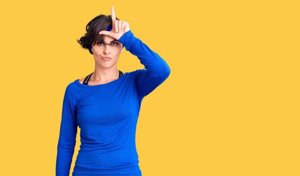 Beautiful young woman with short hair wearing training workout clothes making fun of people with fingers on forehead doing loser gesture mocking and insulting.  - Photo, Image