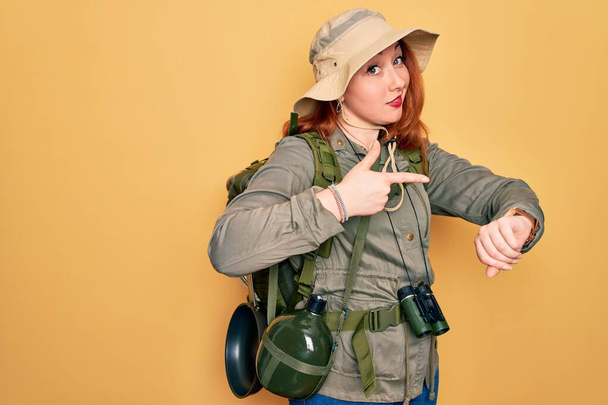 Jonge roodharige backpacker vrouw wandelen dragen rugzak en hoed over gele achtergrond In haast wijzend om tijd te kijken, ongeduld, boos en boos voor deadline vertraging - Foto, afbeelding