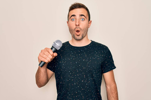 Νεαρός όμορφος τραγουδιστής με μπλε μάτια να τραγουδάει χρησιμοποιώντας μικρόφωνο πάνω από λευκό φόντο φοβισμένος σε κατάσταση σοκ με ένα πρόσωπο έκπληξη, φοβισμένος και ενθουσιασμένος με την έκφραση του φόβου - Φωτογραφία, εικόνα