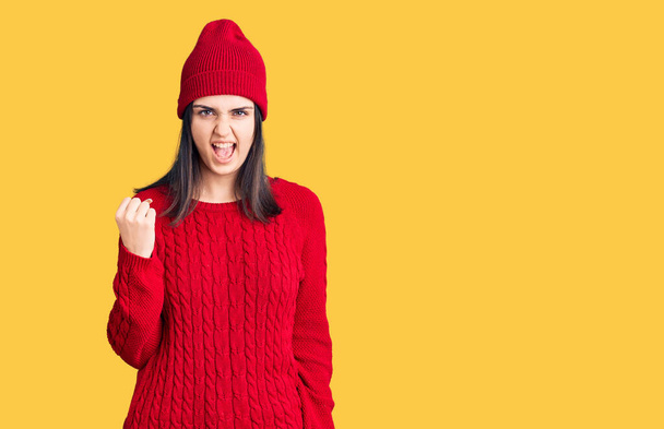 Young beautiful girl wearing sweater and wool cap angry and mad raising fist frustrated and furious while shouting with anger. rage and aggressive concept.  - Photo, Image