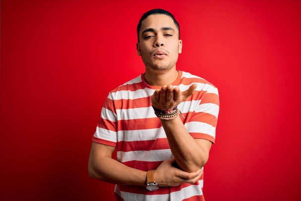 Young brazilian man wearing casual striped t-shirt standing over isolated red background looking at the camera blowing a kiss with hand on air being lovely and sexy. Love expression. - Foto, afbeelding