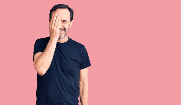 Middle age handsome man wearing casual t-shirt covering one eye with hand, confident smile on face and surprise emotion.  - Photo, Image