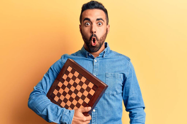 Junger hispanischer Mann hält Schach erschrocken und erstaunt mit offenem Mund für Überraschung, ungläubiges Gesicht  - Foto, Bild