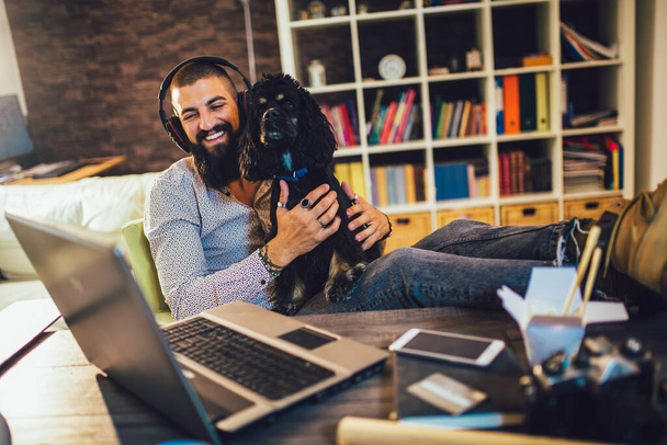 Knappe getatoeëerde man thuis werken op laptop terwijl zitten aan de tafel met schattige hond - Foto, afbeelding