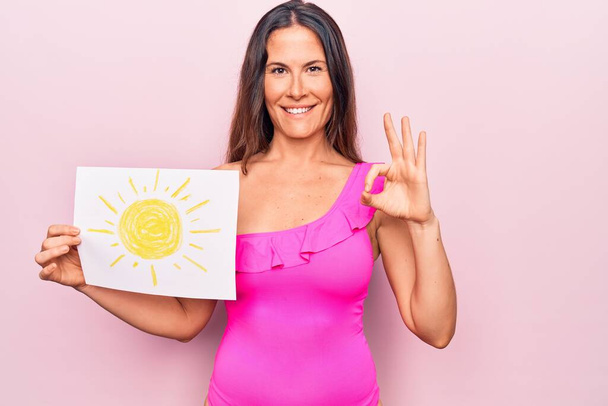 Young beautiful brunette woman wearing pink swimwear holding paper with sun draw doing ok sign with fingers, smiling friendly gesturing excellent symbol - Photo, Image