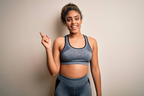 Young african american sportswoman doing sport wearing sportswear over white background with a big smile on face, pointing with hand finger to the side looking at the camera. - 写真・画像