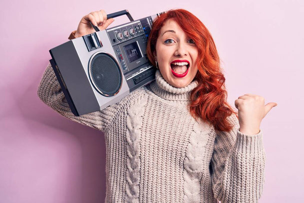 Krásná zrzka žena poslouchá hudbu drží vintage boombox přes růžové pozadí ukazující palec nahoru na stranu usmívá šťastný s otevřenými ústy - Fotografie, Obrázek