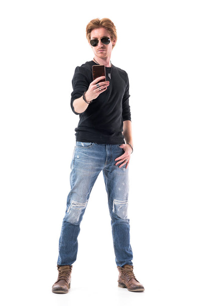 Komea nuori tyylikäs punatukkainen mies farkuissa ottamassa valokuvia matkapuhelimella. Täyspitkä muotokuva eristetty valkoisella pohjalla.  - Valokuva, kuva