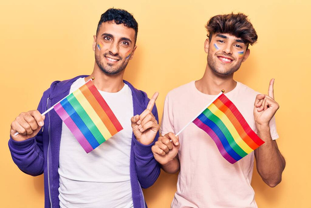 jong gay paar holding regenboog lgbtq vlaggen glimlachen gelukkig wijzend met hand en vinger naar de zijkant  - Foto, afbeelding