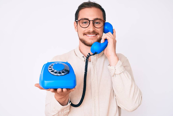 Joven hombre guapo sosteniendo teléfono vintage buscando positivo y feliz de pie y sonriendo con una sonrisa confiada mostrando los dientes  - Foto, imagen