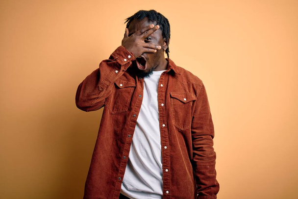 Young handsome african american man wearing casual jacket standing over yellow background peeking in shock covering face and eyes with hand, looking through fingers with embarrassed expression. - Foto, afbeelding