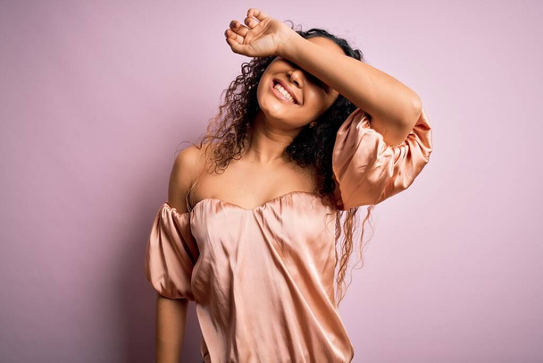 Young beautiful woman with curly hair wearing casual t-shirt standing over pink background covering eyes with arm smiling cheerful and funny. Blind concept. - Fotó, kép