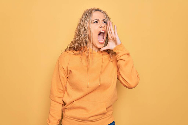 Jonge mooie blonde sportieve vrouw draagt casual sweatshirt over gele achtergrond schreeuwen en schreeuwen luid naar zij met de hand op de mond. Communicatieconcept. - Foto, afbeelding