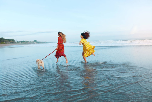 Girls With Dog On Beach. Running Women In Maxi Dresses Along Ocean Shore. Female Models In Trendy Outfit On Summer Vacation. Bohemian Clothing Style For Fashion Look On Dog-Friendly Resort. - Photo, image