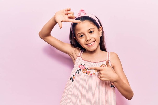 Gyönyörű gyermek lány visel alkalmi ruhák mosolygós így keret kezek és ujjak boldog arcát. kreativitás és fényképezés koncepció.  - Fotó, kép